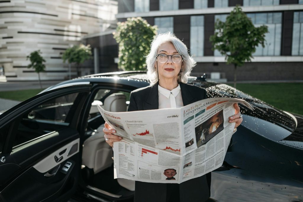 Elderly businesswoman reading a newspaper beside a car outdoors, conveying professionalism and confidence.