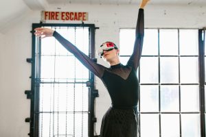 A female ballet dancer in a black outfit using a VR headset, striking a graceful pose indoors.