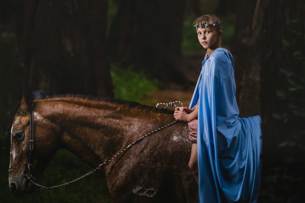 A young child in a blue cloak rides a horse through a mysterious forest setting.