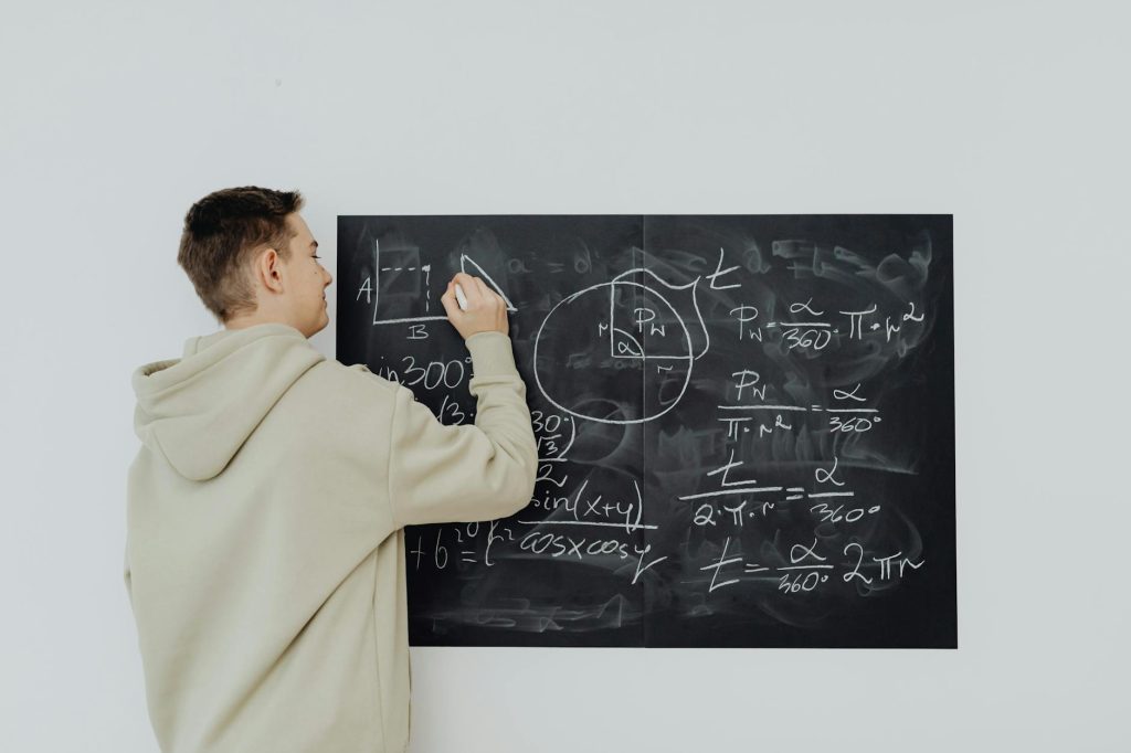 Teen boy in hoodie writes math formulas on a chalkboard, focused on solving algebra equations.