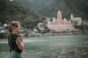 A woman practices yoga by the Ganges River in Rishikesh with a temple in the background.