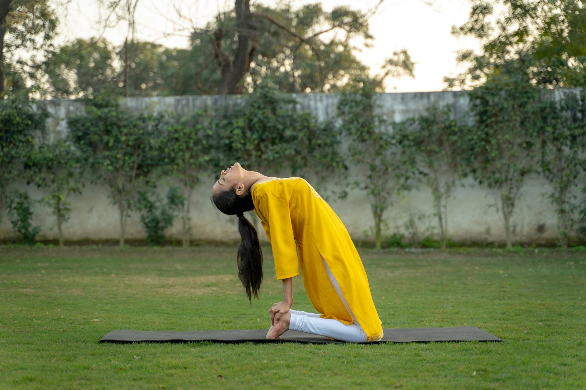 A woman performs a yoga pose in a serene outdoor park, wearing a vibrant yellow traditional outfit.