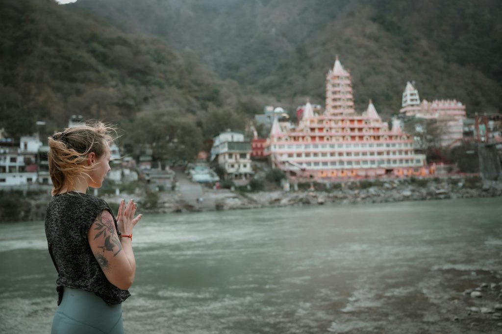 A woman practices yoga by the Ganges River in Rishikesh with a temple in the background.